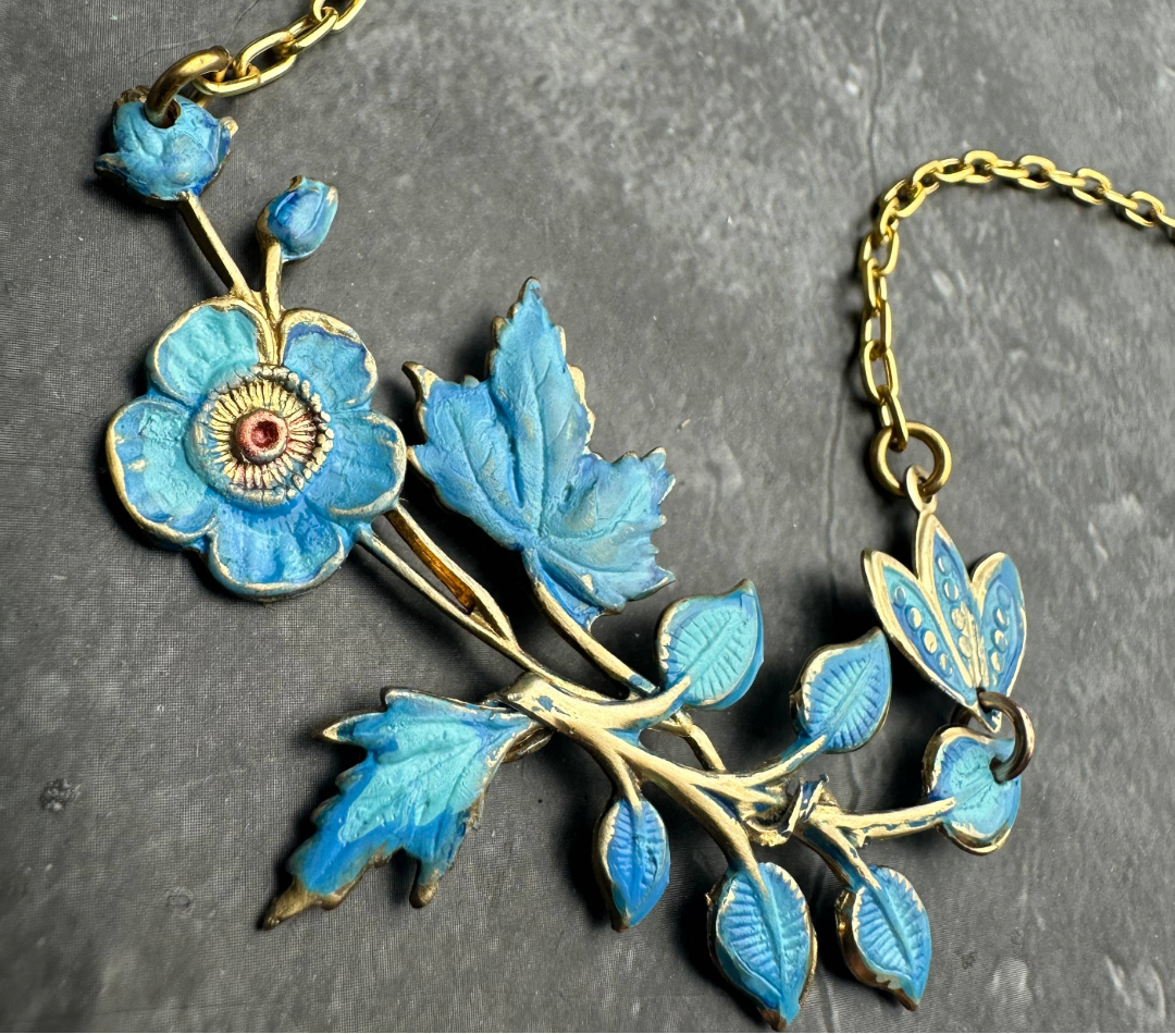 A vintage-style floral pendant necklace with bright blue enamel flowers and leaves on a gold-tone chain, displayed on a dark textured slate surface.